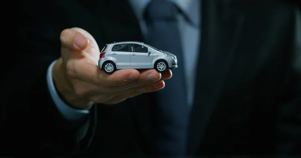 Un hombre vestido con traje y corbata, muestra un pequeño auto. Concepto: seguro de coche, mecánico, arrendamiento, compra de coches nuevos y usados y garajes públicos . — Foto de Stock