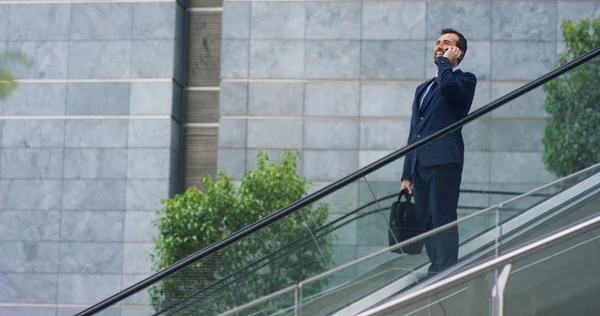 Em escadas rolantes, um homem de negócios atende o telefone, envia mensagens e sorri para a bela notícia de trabalho. Conceito: tecnologia, telefonia, viagens de negócios, negócios . — Fotografia de Stock