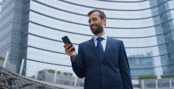Ein Geschäftsmann am Telefon, sendet Nachrichten und lächelt für die schönen Jobnachrichten und im Hintergrund sieht man einen Wolkenkratzer. Konzept: Technologie, Telefonie, Geschäftsreisen, Business, Wall Street — Stockfoto
