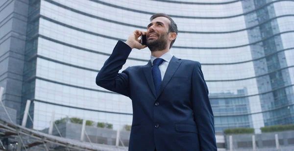 A businessman answering the phone, send messages and smiles for the beautiful job news and in the background you see a skyscraper. Concept: technology, telephony, business trips, business, wall street — Stock Photo, Image