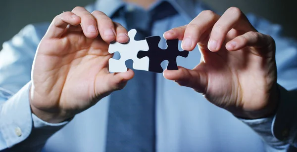 Un hombre de negocios con un traje sosteniendo una pieza de rompecabezas blanco y un negro los une. Concepto: diversidad, trabajo en equipo, racismo y solución de acceso . —  Fotos de Stock