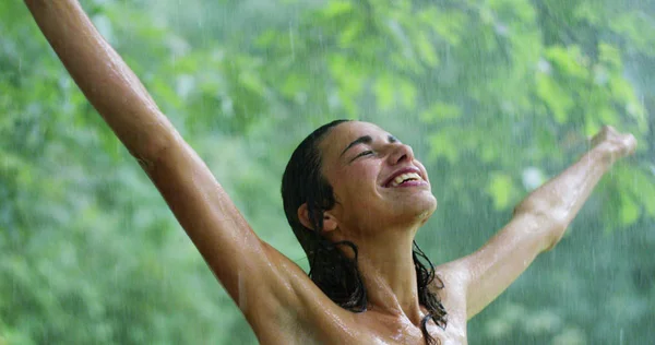 En un caluroso día de verano una mujer sonriendo bajo el agua rodeada de vegetación. Refresca bajo el agua y siente que su cuerpo obtiene el beneficio del agua fresca . — Foto de Stock