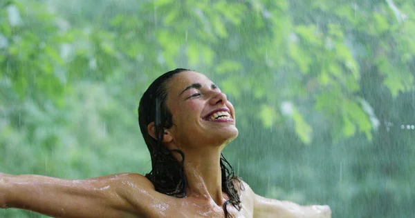 En un caluroso día de verano una mujer sonriendo bajo el agua rodeada de vegetación. Refresca bajo el agua y siente que su cuerpo obtiene el beneficio del agua fresca . — Foto de Stock