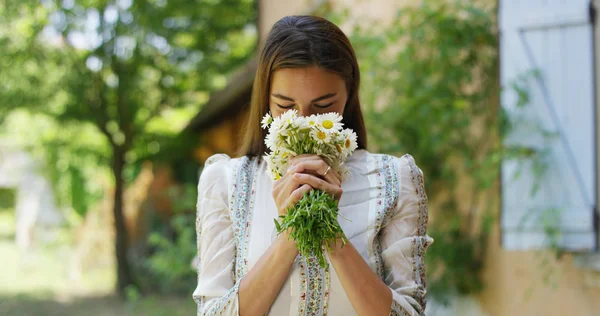 Una mujer hermosa, vestida de blanco puro, huele un ramo de flores de margaritas, rodeada de naturaleza muestra el ramo de flores en la habitación y sonríe. Concepto: amor por la naturaleza, manzanilla, perfumes — Foto de Stock