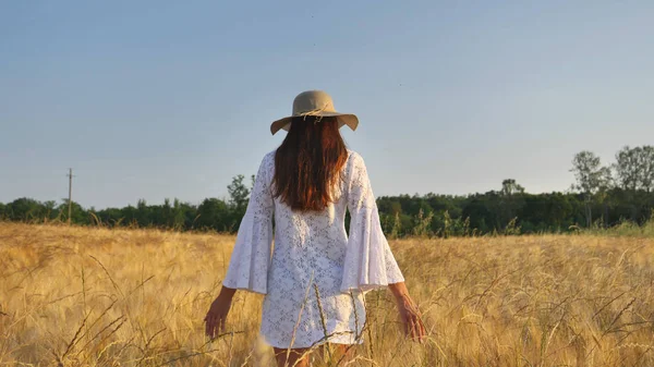 En un día soleado, una mujer está caminando a lo largo de un campo de trigo con un vestido blanco, en un sombrero de paja sobre un fondo de la naturaleza Concepto estilo de vida ecología ambiente feliz gente libertad, maravillosamente hermosa vista — Foto de Stock