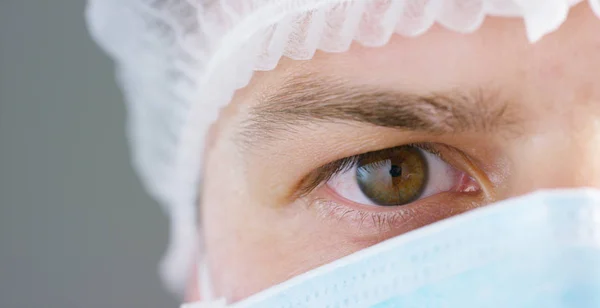 Ett porträtt av en läkare eller kirurg i en medicinsk mask, respirator, bruna ögon, en kirurgisk mössa, i ett sjukhus eller en klinik. Koncept: första medicinsk hjälp, att behandla människor, kirurgi, hälsa, läkare. — Stockfoto