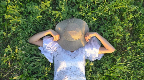 En un día soleado, una mujer en un vestido blanco se encuentra en la hierba verde y esconde su cara con un sombrero de paja Concepto estilo de vida ecología colorido feliz gente libertad maravillosamente hermosa, verdes, medio ambiente — Foto de Stock