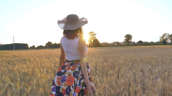 En un día soleado, una mujer está caminando a lo largo de un campo de trigo con un vestido blanco, en un sombrero de paja sobre un fondo de la naturaleza Concepto estilo de vida ecología ambiente feliz gente libertad, maravillosamente hermosa vista — Foto de Stock