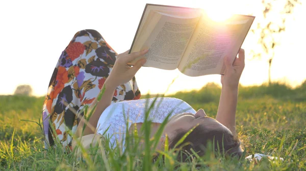 Op een zonnige dag leest een mooie jonge student meisje een boek in de natuur (in een park, op een veld) tegen een achtergrond van gras en hemel. Begrip ecologie, schone lucht, zomer, lente, gras, studie, onderwijs — Stockfoto