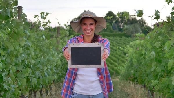 Gadis cantik (wanita) petani tersenyum melihat ladang anggur, memegang papan hitam, mengenakan kemeja, mengenakan topi jerami. Konsep ekologi, anggur bio produk inspeksi air produk alami pertanian — Stok Foto
