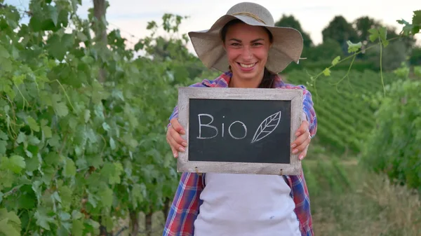 Belle fille (femme) agricultrice souriante regardant les champs de raisin, tenant un tableau noir, dans une chemise, portant un chapeau de paille. Concept écologie, vin bio inspection des produits eau produits naturels agriculture — Photo