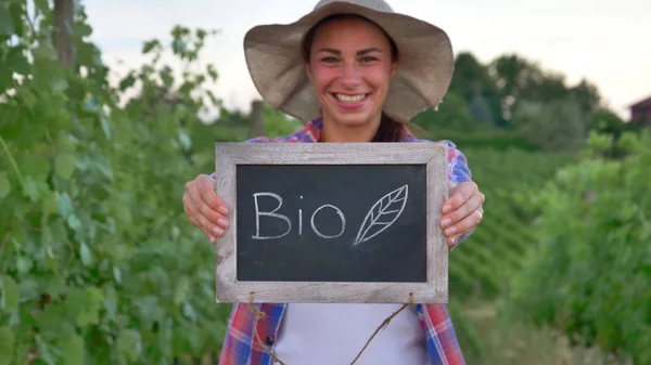 Üzüm alanları izlerken gülümseyen güzel kız (kadın) çiftçi holding siyah bir yönetim kurulu, bir gömlek bir hasır şapka giyiyor. Kavram ekoloji, şarap bio ürün muayene su doğal ürünler tarım — Stok fotoğraf