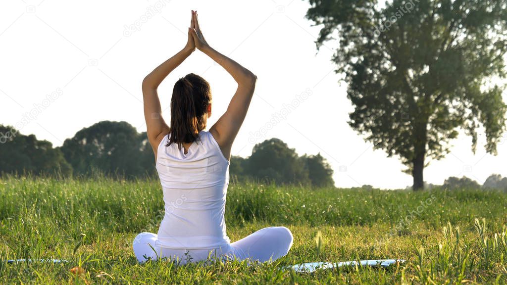 On a sunny day, a beautiful young girl does yoga in nature (park, field), in white clothes in the background of grass, wood, sky. Concept: ecology, clean air, summer, spring, qi energy, harmony.