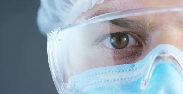 Um retrato de um médico ou cirurgião em uma máscara médica, um respirador, olhos castanhos, um boné cirúrgico, em um hospital ou uma clínica . — Fotografia de Stock