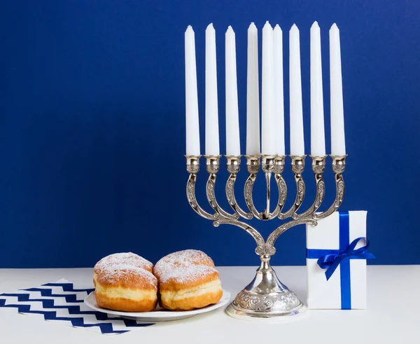 Celebración de Hanukkah. Candelabro con rosquillas fritas, regalo, sobre fondo blanco y azul —  Fotos de Stock