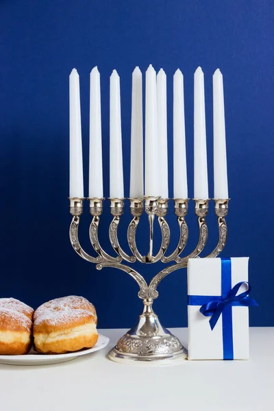Celebración de Hanukkah. Candelabro con rosquillas fritas, regalo, sobre fondo blanco y azul —  Fotos de Stock