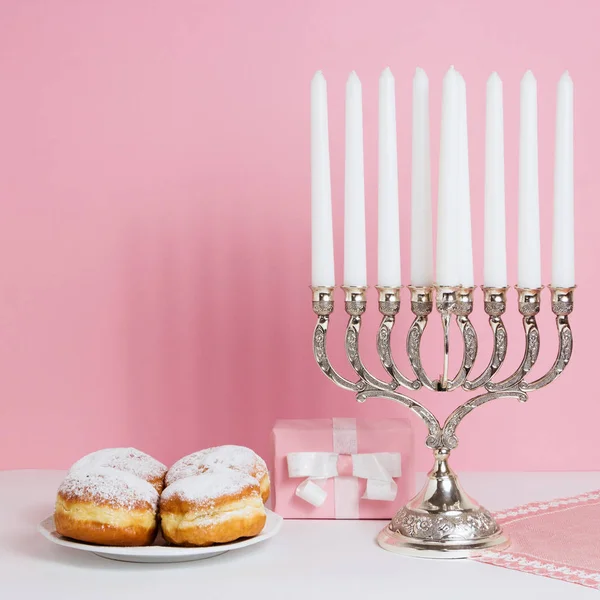 Celebración de Hanukkah. Candelabro con rosquillas fritas, regalo, sobre fondo blanco y rosa —  Fotos de Stock
