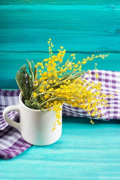 Flores amarelas da primavera de mimosa em uma caneca branca em um fundo de madeira azul . — Fotografia de Stock