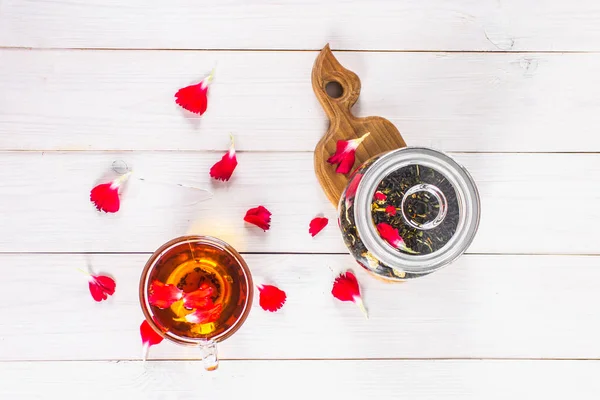 Una tazza di tè, petali e un barattolo con tisana floreale nera su un tavolo di legno bianco. Vista dall'alto . — Foto Stock