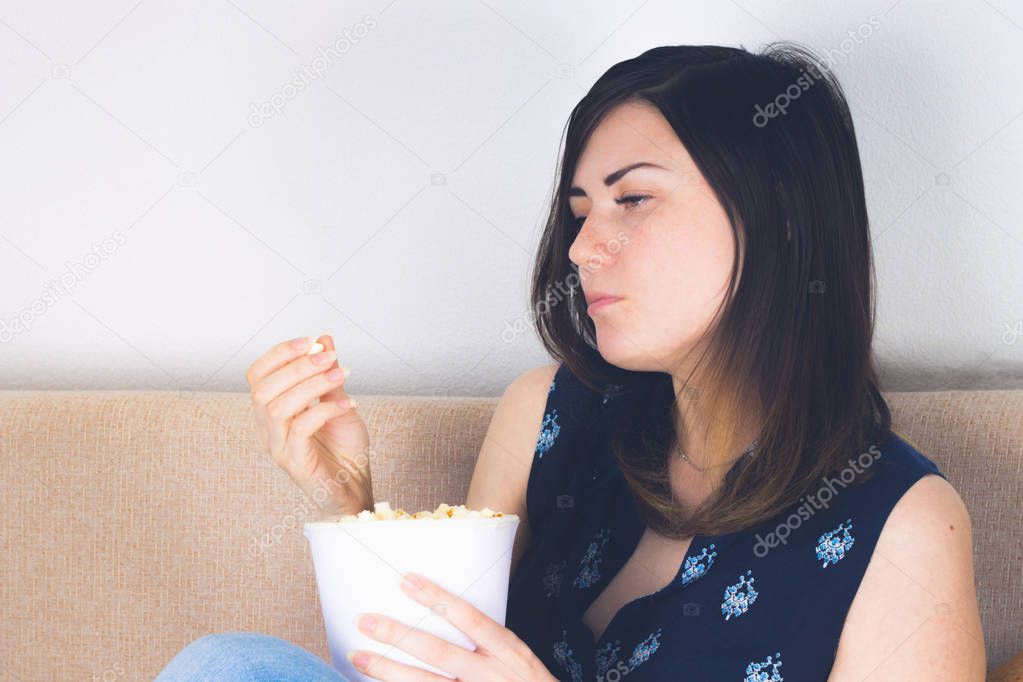 A girl on the couch watching TV and eating popcorn. Emotions are sadness, depression.