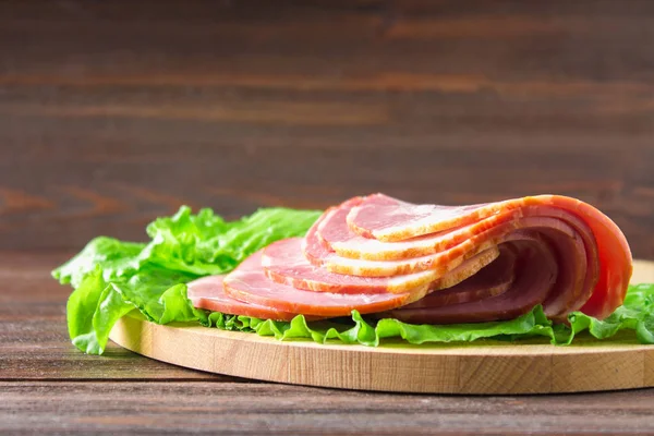 Taze yeşil salata ile dilimlenmiş jambon yuvarlak kesme tahtası üzerinde bırakır. Et ürünleri bir kahverengi ahşap tablo.