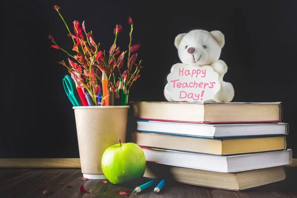 Concept of Teacher's Day. Objects on a chalkboard background. Books, green apple, bear with a sign: Happy Teacher's Day, pencils and pens in a glass, twig with autumn leaves.