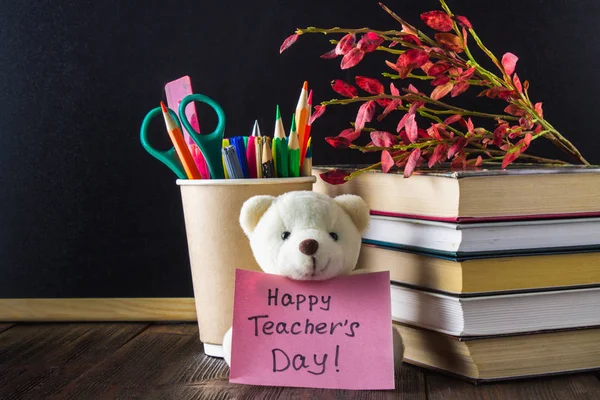 Concept of Teacher's Day. Objects on a chalkboard background. Books, green apple, bear with a sign: Happy Teacher's Day, pencils and pens in a glass, twig with autumn leaves.