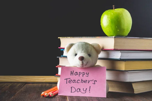 Konzept des Lehrertages. Objekte auf einem Tafelhintergrund. Bücher, grüner Apfel, Bär mit Schild: fröhlicher Lehrertag, Bleistifte und Stifte im Glas, Zweige mit Herbstblättern. — Stockfoto