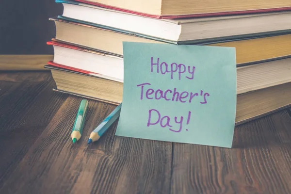 Concept of Teacher\'s Day. Objects on a chalkboard background. Books, green apple, plaque: Happy Teacher\'s Day, pencils and pens in a glass, sprig with autumn leaves.