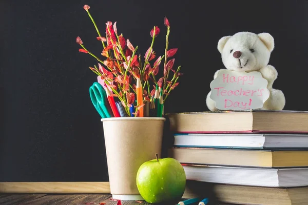 Concept of Teacher\'s Day. Objects on a chalkboard background. Books, green apple, bear with a sign: Happy Teacher\'s Day, pencils and pens in a glass, twig with autumn leaves.