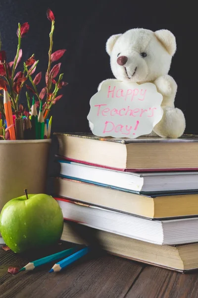 Concept of Teacher's Day. Objects on a chalkboard background. Books, green apple, bear with a sign: Happy Teacher's Day, pencils and pens in a glass, twig with autumn leaves.