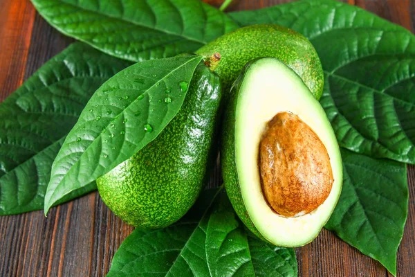 Drei grüne rohe reife Avocadofrüchte und eine geschnittene Hälfte mit Knochen und Blättern auf einem braunen Holztisch. — Stockfoto