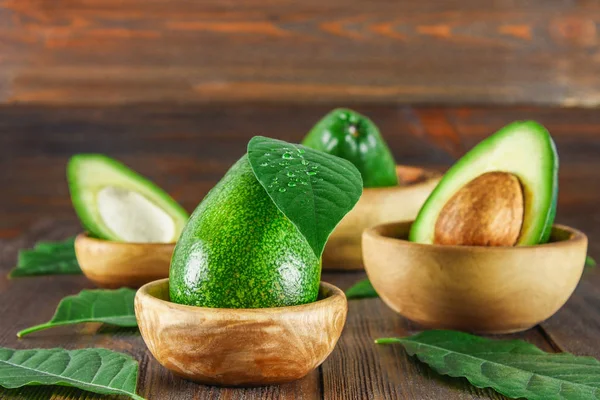 Grüne rohe reife Schnitt- und ganze Avocadofrüchte mit Stein liegen in hölzernen Schalen umgeben von Blättern auf einem braunen Tisch. — Stockfoto
