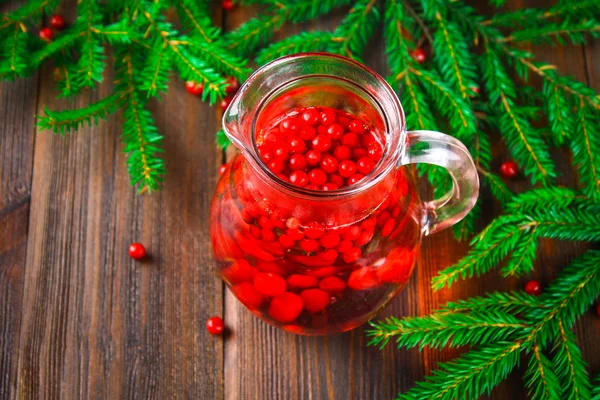 Morse ou chá de cowberry em um jarro de vidro cercado por ramos de abeto em uma mesa de madeira . — Fotografia de Stock