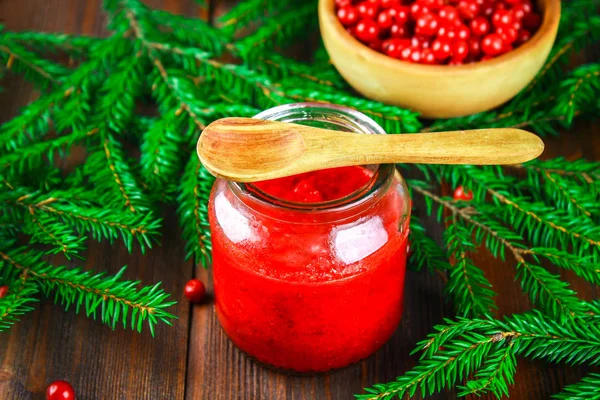 Cowberry jam in a glass jar on a wooden table surrounded by berries and fir branches. — Stock Photo, Image