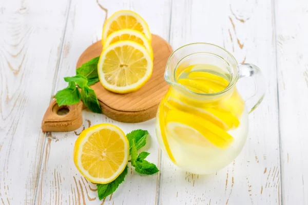 Um jarro com uma limonada fria em um fundo de madeira branco cercado por limões . — Fotografia de Stock
