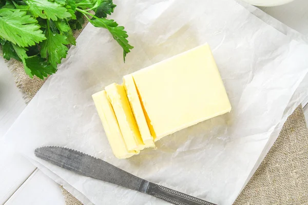 Una barra de mantequilla se corta en trozos en una tabla de madera con un cuchillo, rodeado de leche, huevos y perejil en una mesa blanca. Ingredientes para cocinar . — Foto de Stock