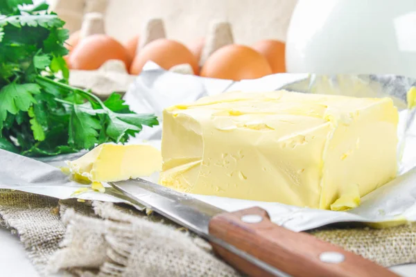 Une barre de beurre est coupée en morceaux sur une planche de bois avec un couteau, entourée de lait, d'œufs et de persil sur une table blanche. Ingrédients pour la cuisine . — Photo