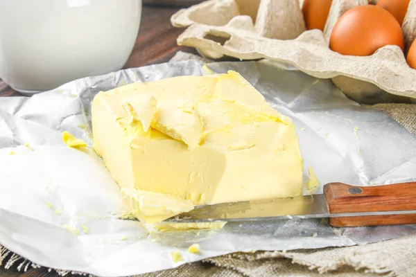 Una barra de mantequilla se corta en trozos en una tabla de madera con un cuchillo, rodeado de leche, huevos y perejil en una mesa marrón. Ingredientes para cocinar . — Foto de Stock