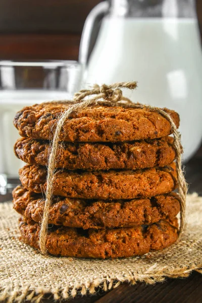 Homemade oatmeal cookies. A stack of cookies tied with string on sackcloth on a brown wooden table. Milk in the background. — Stock Photo, Image