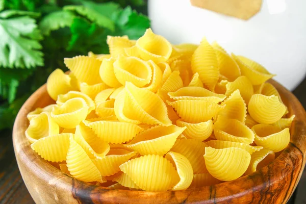 Fondo culinario con pasta conchiglie sobre mesa de madera. Pasta en forma de conchas en un tazón de madera con perejil . — Foto de Stock