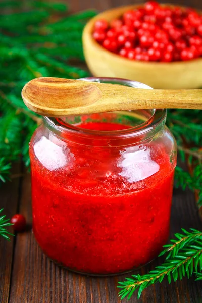 Preiselbeermarmelade im Glas auf einem Holztisch, umgeben von Beeren und Tannenzweigen. — Stockfoto