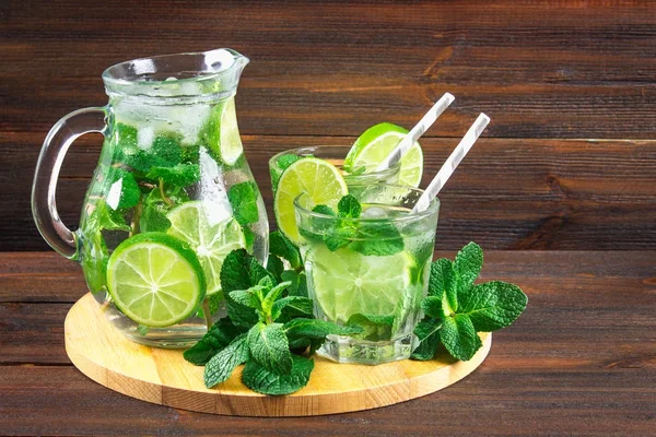 Mojito com hortelã e limão em um copo e um jarro na tábua redonda. Fundo de madeira marrom — Fotografia de Stock