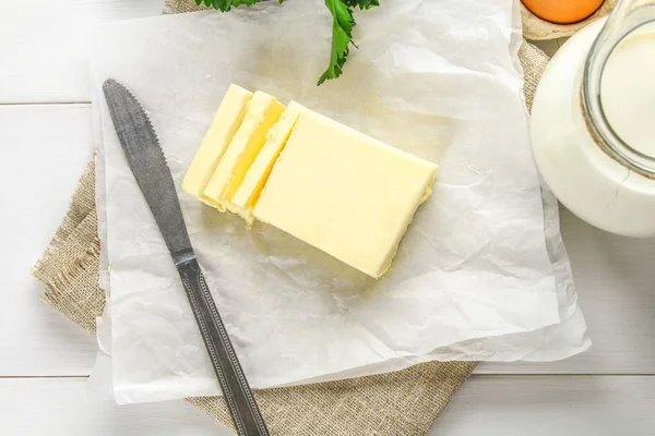 Una barra de mantequilla se corta en trozos en una tabla de madera con un cuchillo, rodeado de leche, huevos y perejil en una mesa blanca. Ingredientes para cocinar . — Foto de Stock