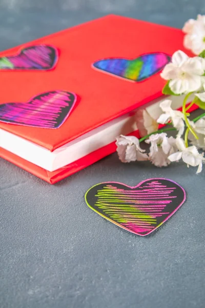 Black and pink hearts on a book in a red cover and flowers on a gray concrete background. The concept of Valentine\'s Day. A symbol of love.