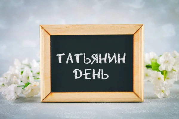 The inscription in Russian: Tatyanin day. Russian holiday on student\'s day. A chalkboard is surrounded by white flowers on a gray background.