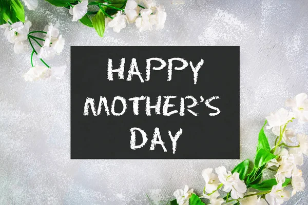 Feliz día de las madres. Una pizarra está rodeada de flores blancas sobre un fondo gris . — Foto de Stock