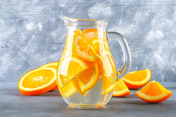 Orange detox water in a pitcher on a gray concrete background. Healthy food, drinks.