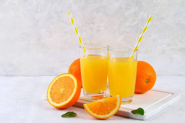 Glass cups and a pitcher of fresh orange juice with slices of orange and yellow tubes on a light gray table. — Stock Photo, Image