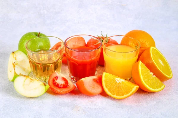 Vasos con naranja fresca, manzana, jugo de tomate sobre una mesa de hormigón gris. Lobulillos Frutas y verduras alrededor . — Foto de Stock